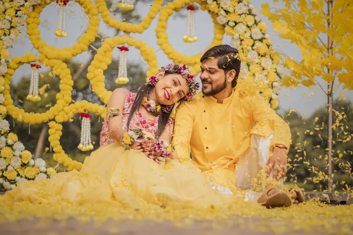 Couple seated together during the haldi, both in yellow, marigolds behind them