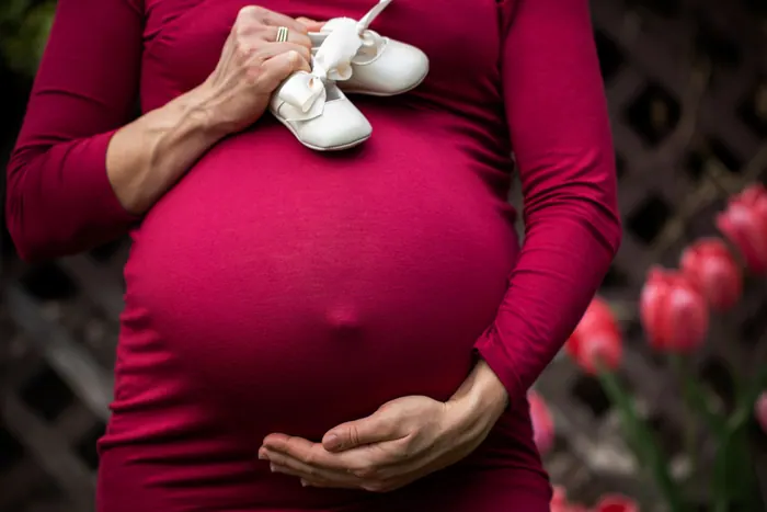 Expectant mother in a deep pink drape holding a pair of baby shoes