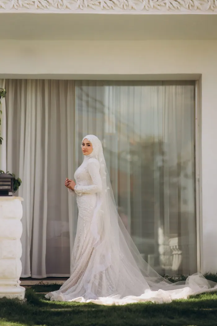 Bride in a white hijab and veil standing by a window before the ceremony
