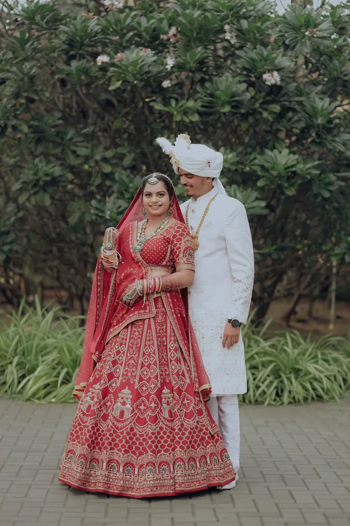 Couple standing together in a garden, the bride in a red lehenga and the groom in a cream sherwani